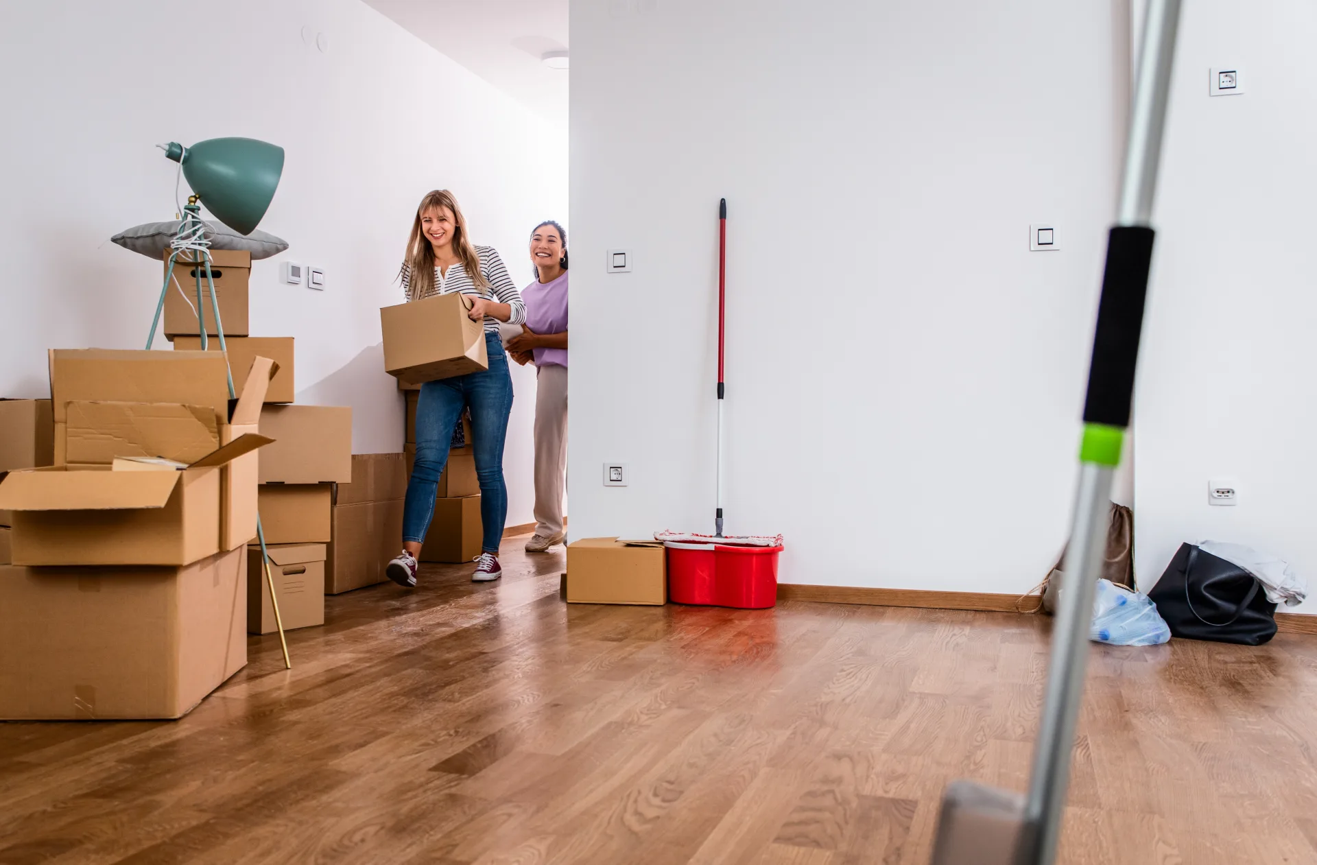 An empty room with a wood floor, a mop and bucket by the wall, and two people carrying boxes in